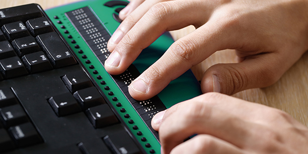 Neben einer Computertastatur befndet sich eine Brailletastatur, auf der jemand liest.