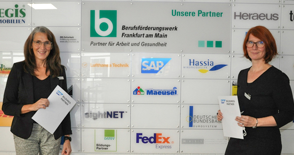 Zwei Frauen halten jeweils ein Zertifikat in der Hand und stehen vor einer Wand, welche die Logos der Parntner des BFW-Frankfurt abbildet. 