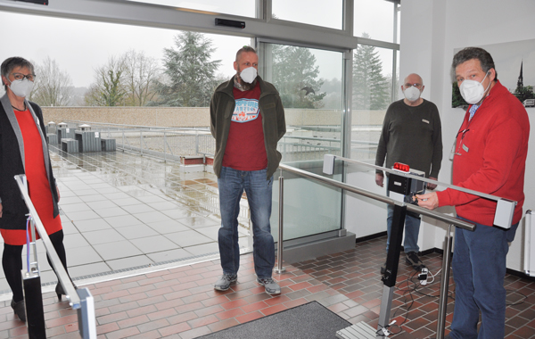 Vier Personen mit Mund-Nasen-Schutz stehen in einer Eingangshalle mit dem Rücken zu einer offenen Schiebetür und schauen in die Kamera.