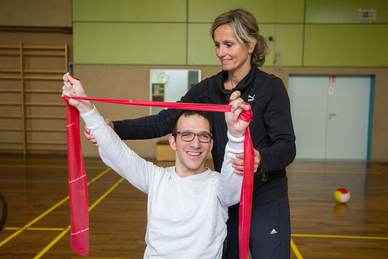 In einer Sporthalle macht eine Frau mit einem Teilnehmer eine Fitnessübung mit einem Band.