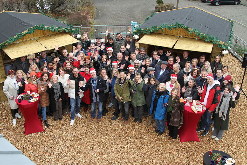 Gruppenfoto vor zwei Holzhütten im weihnachtlich gestalteten Außenbereich. Manche tragen Weihnachtsmützen, einige winken in die Kamera.