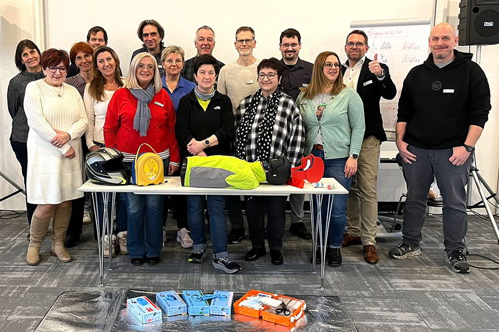 Gruppenfoto der Teilnehmenden des Erste-Hilfe-Auffrischungskurses. Auf einem Tisch und auf dem Boden liegen verschiedene Hilfsmittel wie z. B. eine Reanimationspuppe und ein Erste-Hilfe-Kasten.