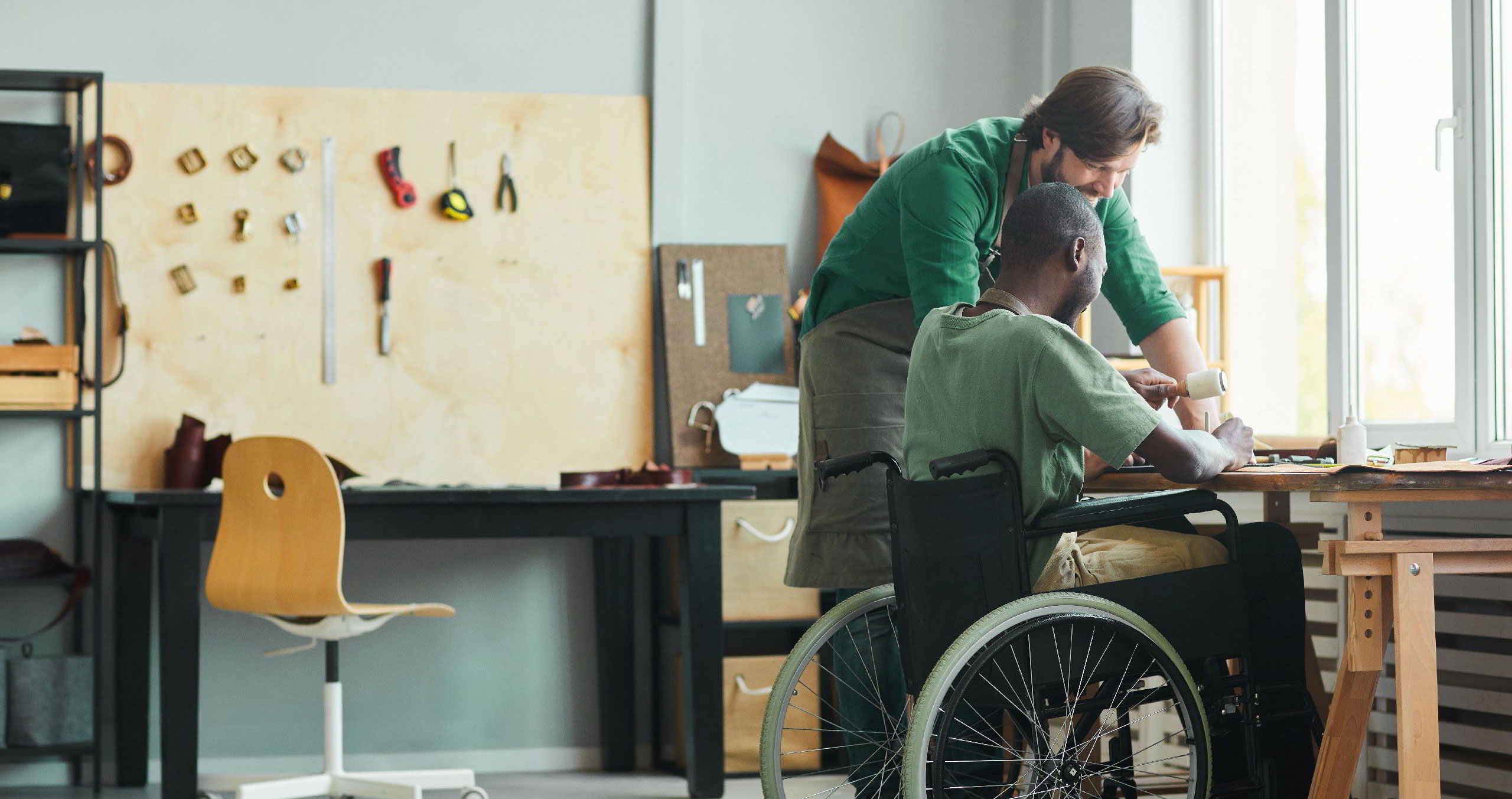 Blick in einen Werkraum. Ein Mann erklärt einem Teilnehmer im Rollstuhl etwas während er an einem Werkstück arbeitet.
