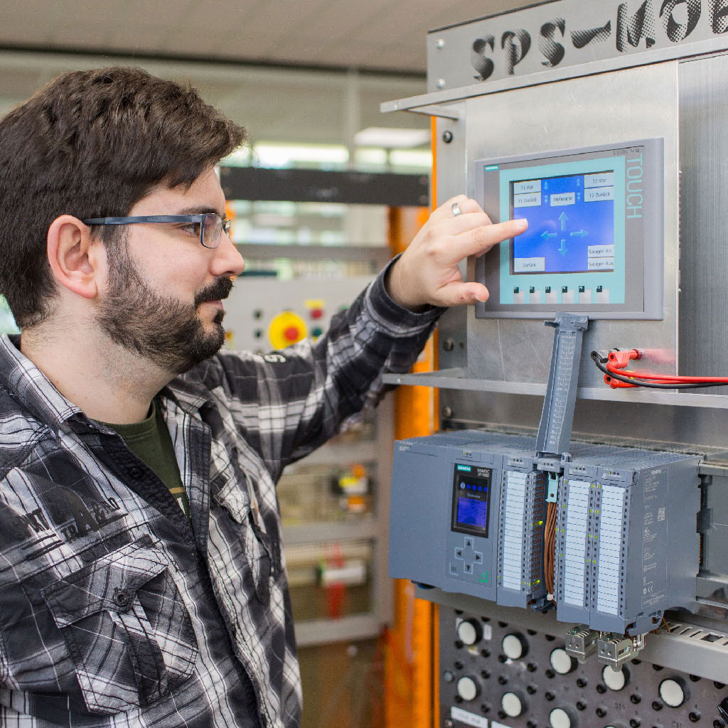 Ein junger Mann mit Bart bedient einen Touchscreen an einem industriellen Schaltschrank mit Steuerungseinheit in einer Art Werkhalle