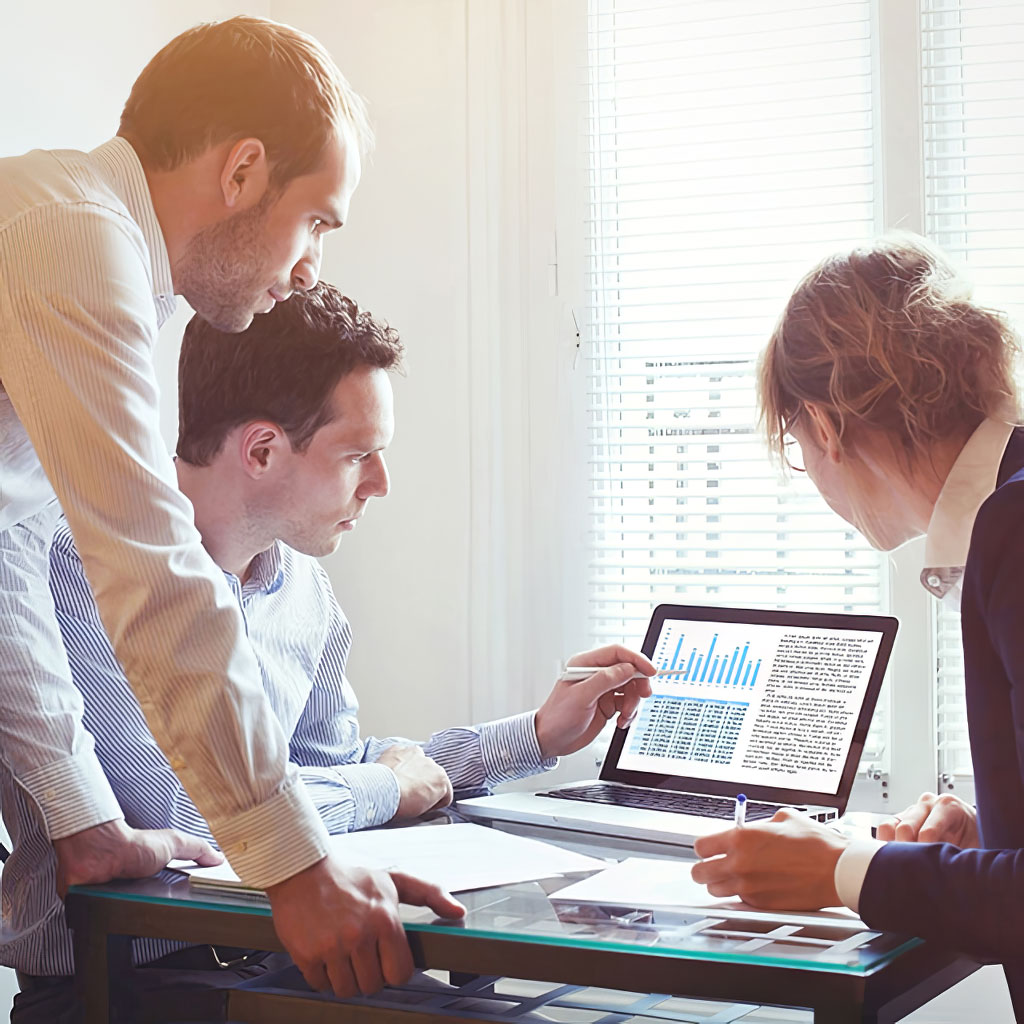 Drei Personen (zwei Männer und eine Frau) besprechen gemeinsam Diagramme und Tabellen auf einem Laptop in einem Büro