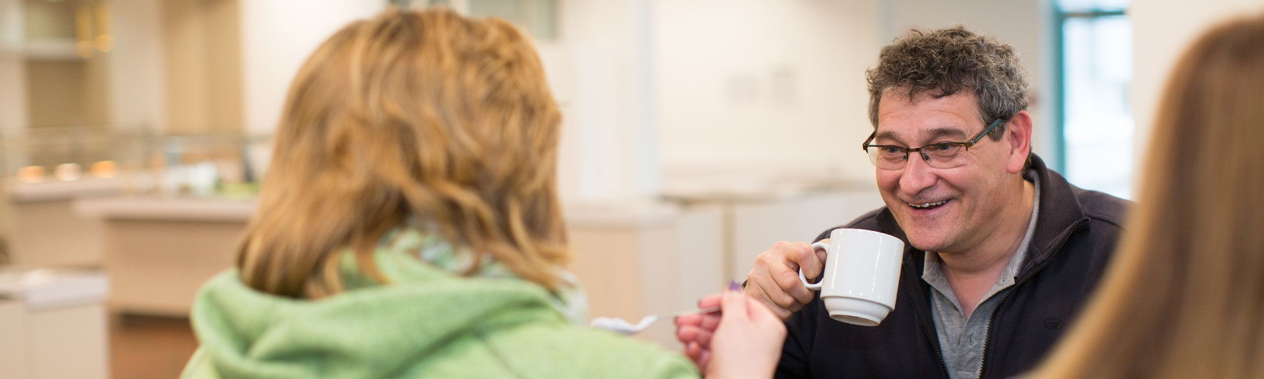 Teilnehmer sitzen mit zusammen beim Essen und unterhalten sich, ein Mann hält eine Tasse und lächelt sein Gegenüber an..  